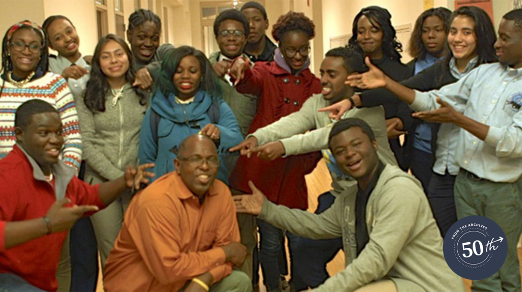 Cosby Hunt kneeling in the front row of a group shot of students, all pointing at him and smiling.