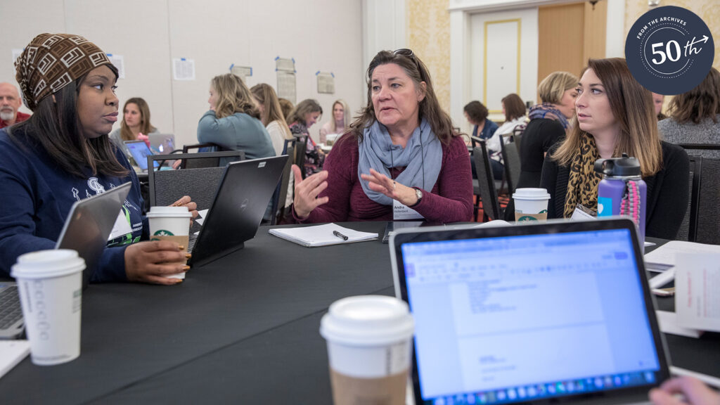 Three teachers having a discussion at a conference table with cups of coffee and a laptop in the foreground.