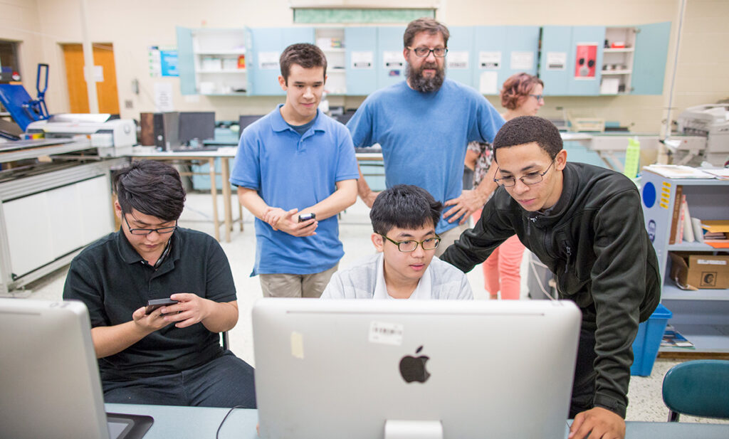Front view of high-school student working on iMac with two others plus teacher looking on.