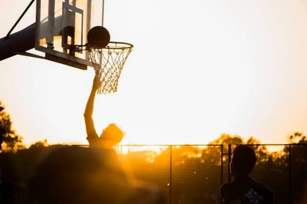 Person doing a layup on an outdoor basketball court, silhouetted by the sunset.