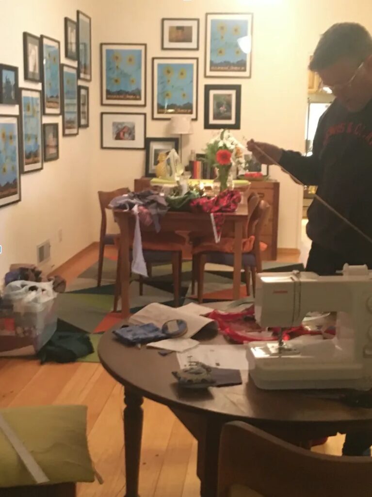 Man standing with tape measure in dining room, framed images on wall.