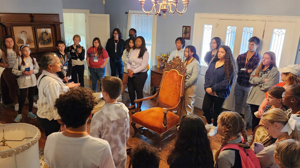 Group of youth at historic site gathered around antique chair in middle of room.