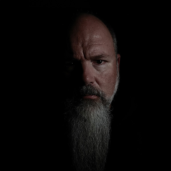 Headshot of Brian Johnson with shadow covering most of left face, black background.